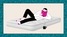 A person relaxing on a mattress, reading a book with a steaming mug nearby.