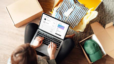 A person sits on the floor using a laptop to shop online, surrounded by open boxes with clothing items including a striped shirt and a green cap.