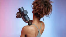 A woman in fitness attire using a massage gun on her shoulder.