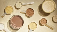 Various scoops of protein powder in different colors, along with glasses of chocolate and vanilla protein shakes, arranged on a light-colored surface.