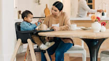 parent feeding baby in UPPAbaby Ciro High chair at dining room table with parent using kitchen faucet in background