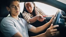 A teen sitting in the driver's seat of a car with an adult reaching in through the driver's side window and pointing at something.