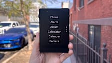 handing hold Light Phone III with navigation menu on the screen, cars and brick building in the background