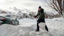 person shoveling deep snow from driveway in suburban neighborhood with other houses in background