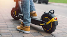 A close-up shot of a person's lower legs and feet while riding a black electric scooter on a paved cobblestone sidewalk.