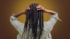 Back view of a person with long, thin braids holding their hands on top of their head against a plain golden-brown background.
