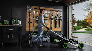 person pushing Ego 56V self propelled lawn mower out of garage with wall of other Ego lawn equipment hanging on garage wall behind them