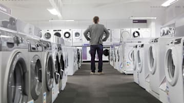 back of person wearing jeans and blazer with hands on hips, standing in row of washer and dryers and looking at wall of washers and dryers