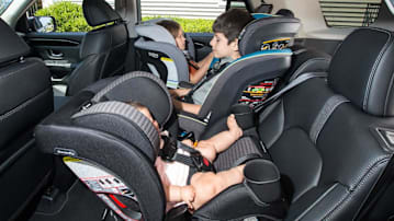 A side view shows three children secured in car seats arranged side-by-side across a vehicle'a back bench seat. In the foreground, an infant lies in a rear-facing car seat, while a young boy sits in a forward-facing car seat in the middle. On the far side, a young girl is secured in a rear-facing car seat.
