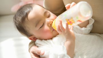 Baby in a caregiver's arms holding and drinking from a bottle.