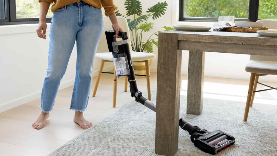 a woman using a Shark Stratos Cordless Stick Vacuum IZ862H to clean a rug under a dining table