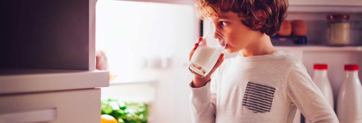 Little boy in his pajamas having a bedtime snack, at nighttime. He is standing next to opened refrigerator, with a glass of milk in his hands. Home interior shot.