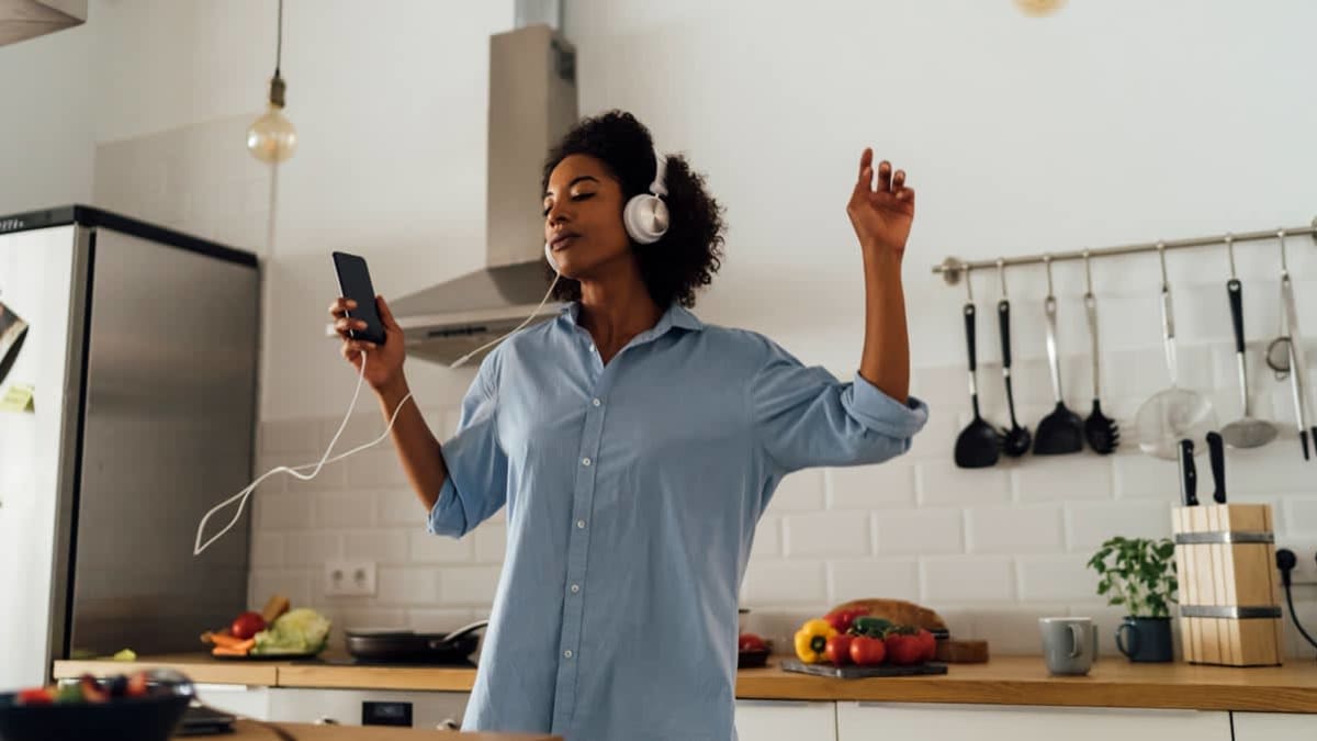 A person in a kitchen wearing a pair of wired headphones while holding a smartphone