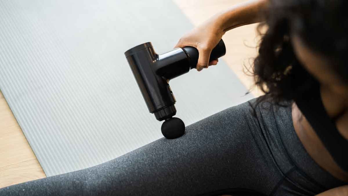 person using massage gun on thigh while sitting on yoga mat