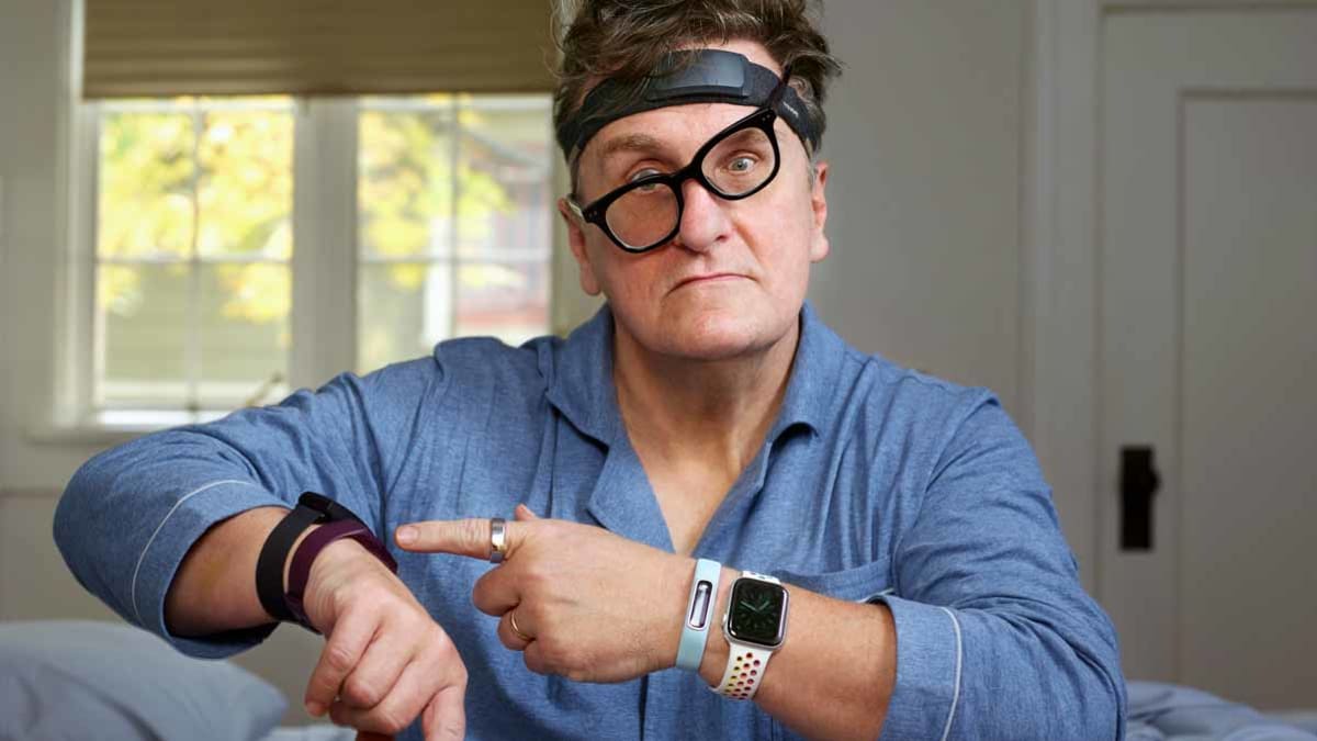person sitting on edge of edge of bed wearing 6 sleep trackers and pointing to sleep tracker