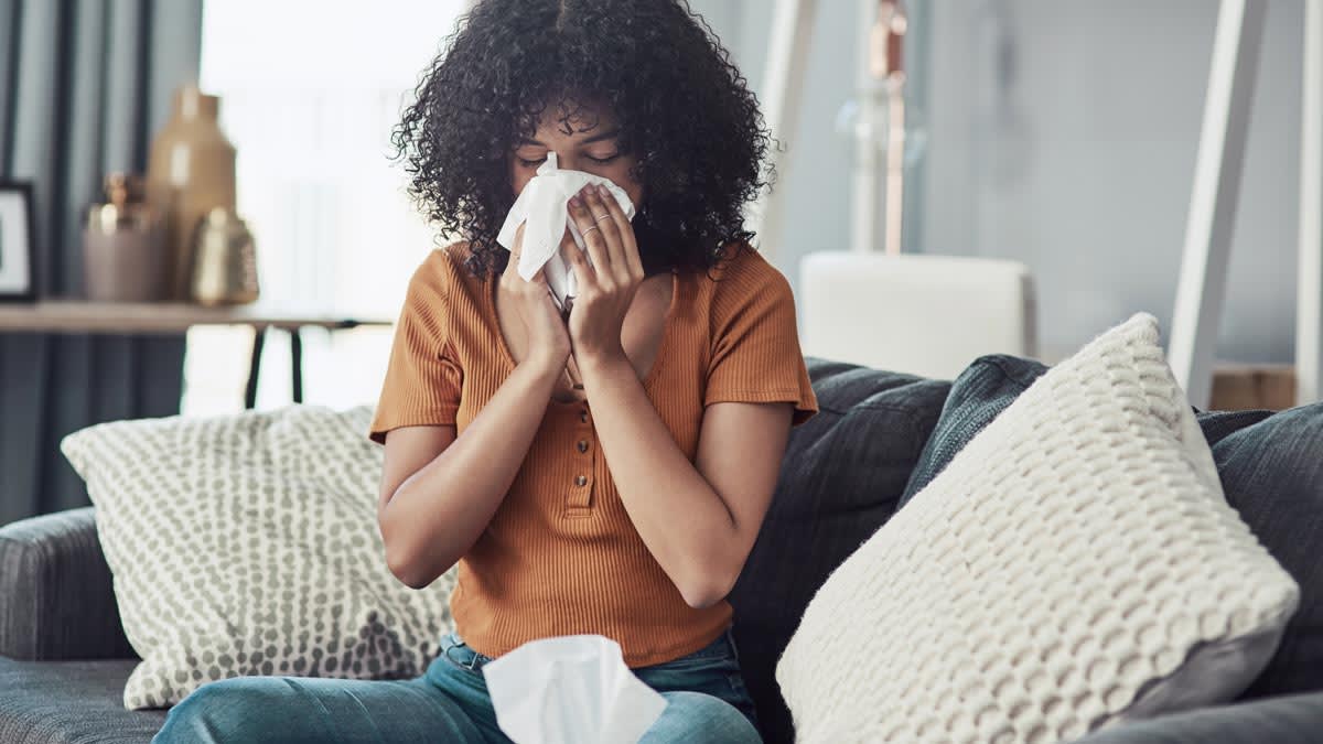 person using tissue to blow nose while sitting on grey couch with 2 large throw pillows