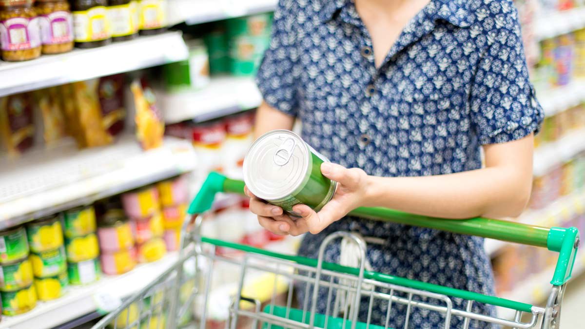 detail of person in grocery store aisle standing in front of grocery cart holding canned food