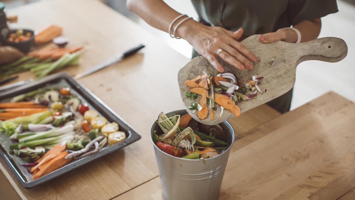 detail of person putting veggie scrapes from wooden cutting board into compost bin with veggies on cooking tray in background