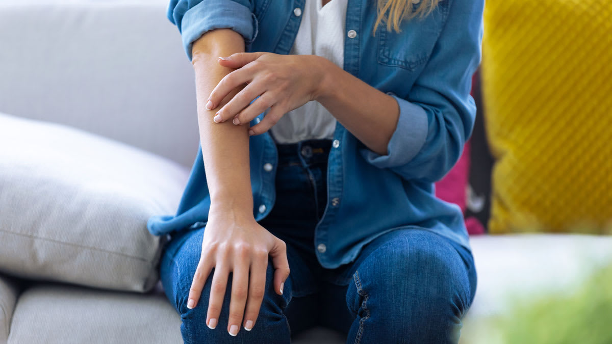 crop of person sitting on couch scratching right arm, wearing denim shirt and jeans