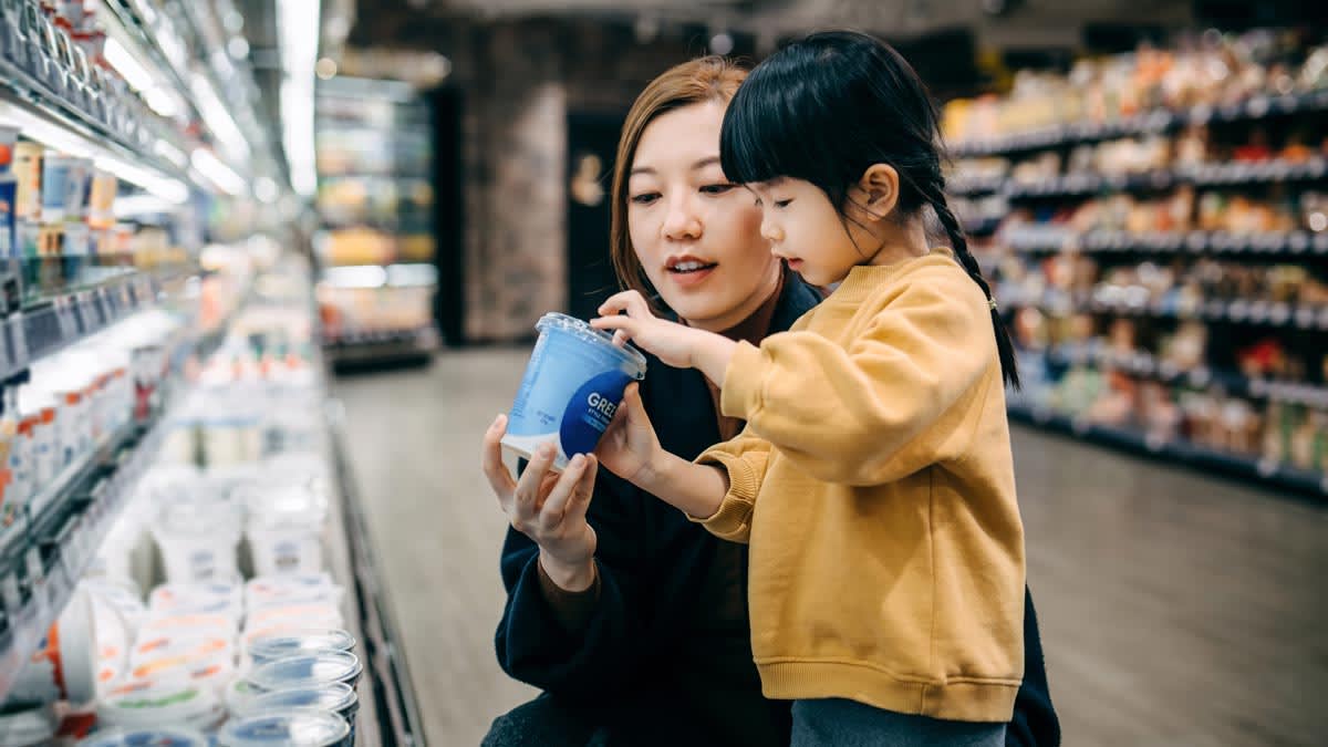 adult showing container of greek yogurt to child in grocery store aisle