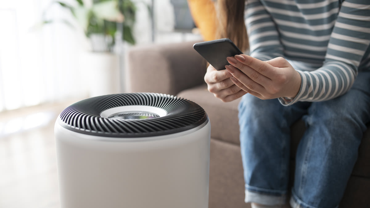 detail of air purifier and person sitting on couch using smartphone to program the air purifier