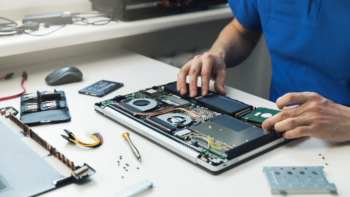 detail of person at table repairing laptop, tools and computer parts surrounding the laptop