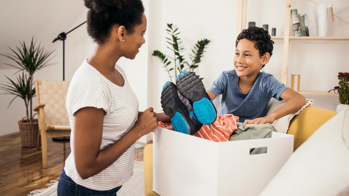 parent and child putting donation items into box in their living room, plants and chair behind them
