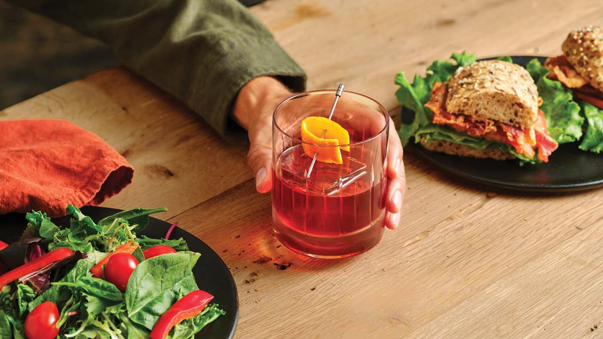 Hand holding a cocktail glass with an orange peel, and plates with salad and sandwiches on a wooden table.