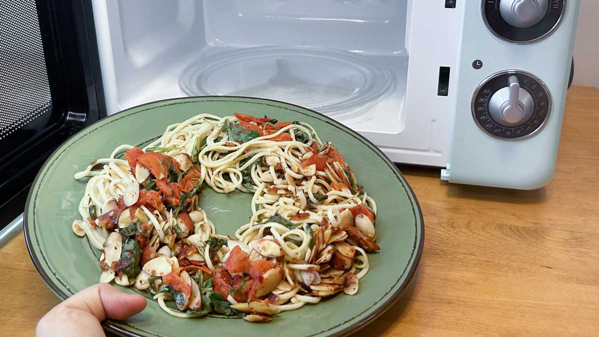 hand holding a plate of pasta with tomato, onion, nuts, and greens made into an open circle on green plate in front of a microwave