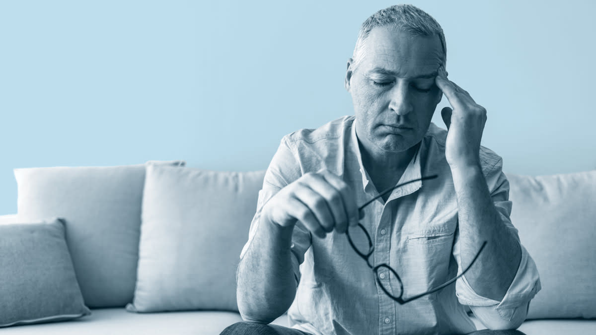 person on couch with eyes closed, holding a pair of glasses in one hand with their other hand on their temple