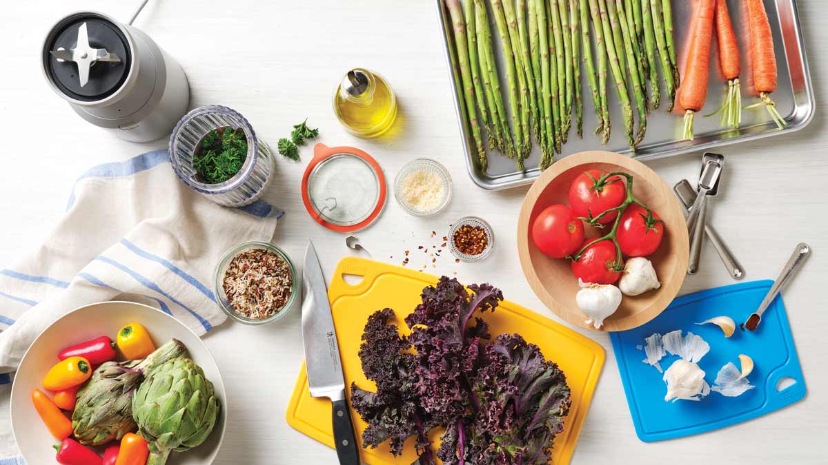 overhead view with Beast 10 Personal Blender, Made in Cookware Half Sheet pan with carrots and asparagus, Luflicon Silicone Cutting board in yellow and blue with kale and garlic, chef's knife, and bowl of peppers and artichokes