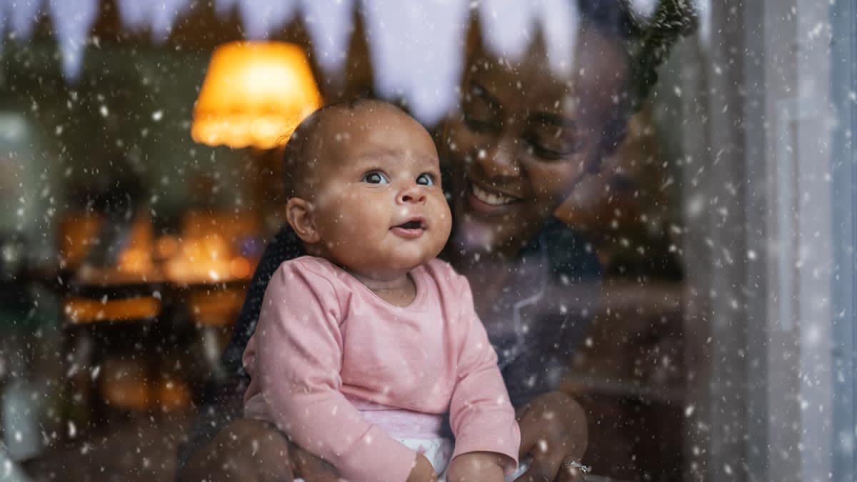 A baby and her mother sitting inside looking at snow falling through a window