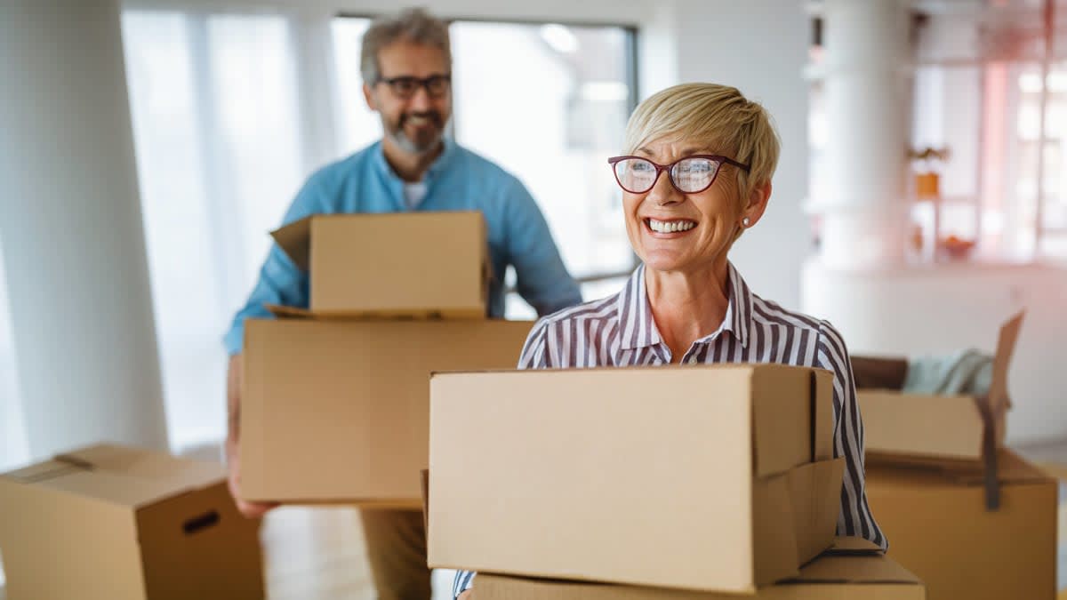 two people carrying cardboard boxes in room with other boxes
