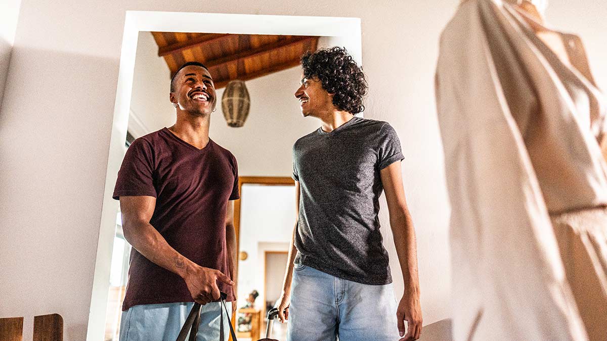 Happy gay couple in summer clothes arriving with their bags at a house.