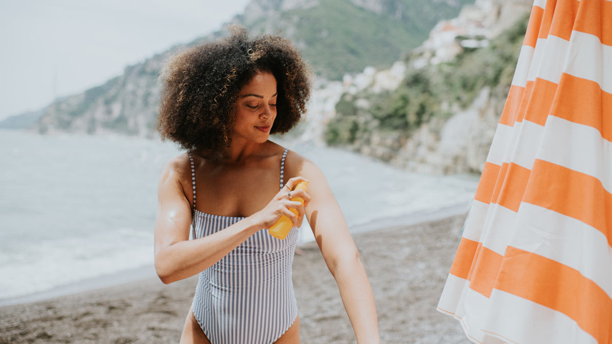person in bathing suit applying spray sunblock on beach with water, beach umbrella, and mountains in background