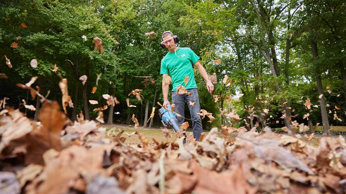 person wearing green Consumer Reports shirt while using leaf blower, leaves in foreground and trees in background