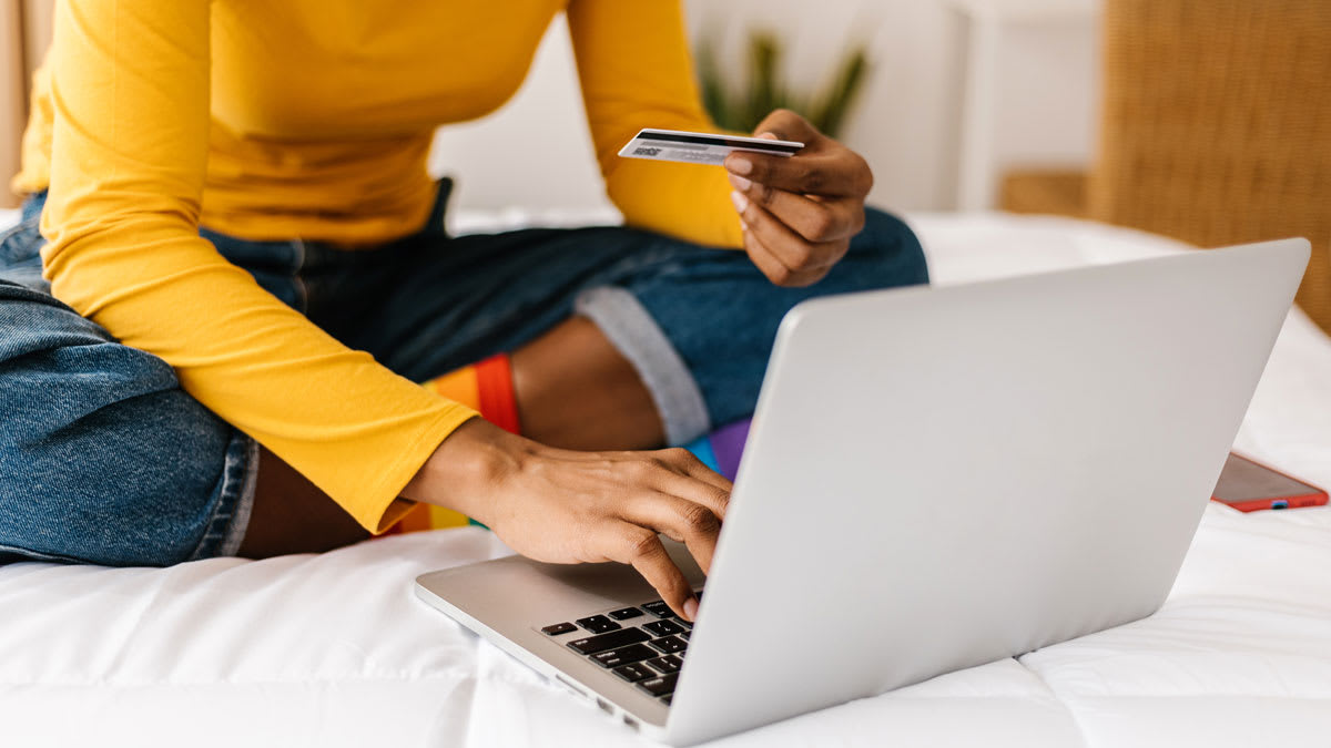 close up of person sitting cross legged on a bed while holding credit card and using a laptop