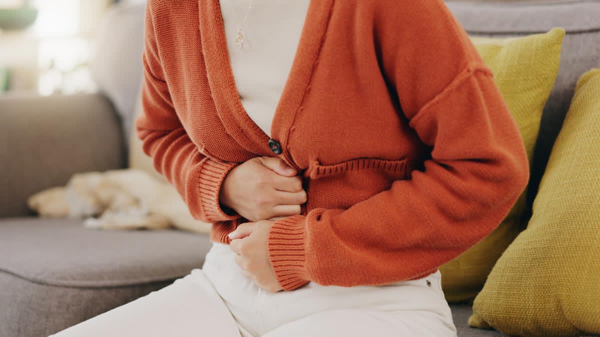 close up of person sitting on couch wearing orange sweater, hands over stomach as though they're in pain