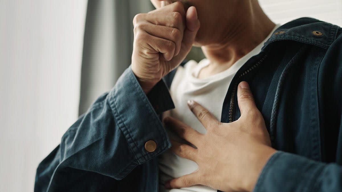 Closeup of person coughing with one hand over their mouth and the other on their chest.