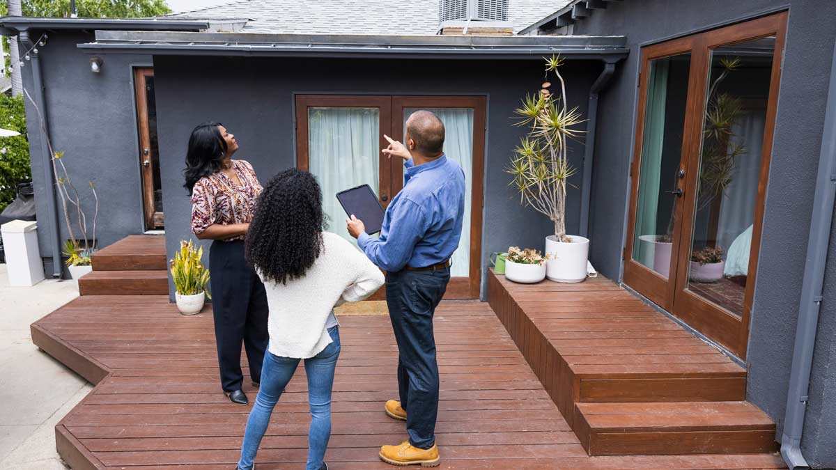 couple looking at home with home inspector holding tablet, pointing to roof of house while standing on wooden deck