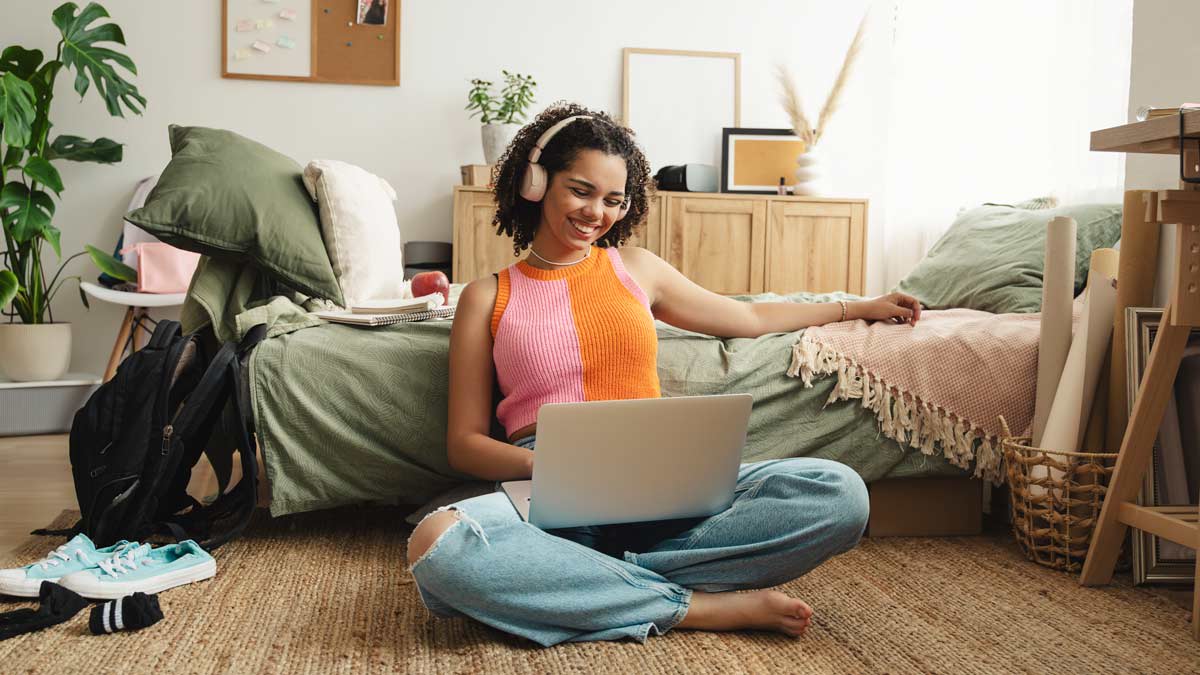 A college student sitting in a dorm room with a laptop and headphones on.