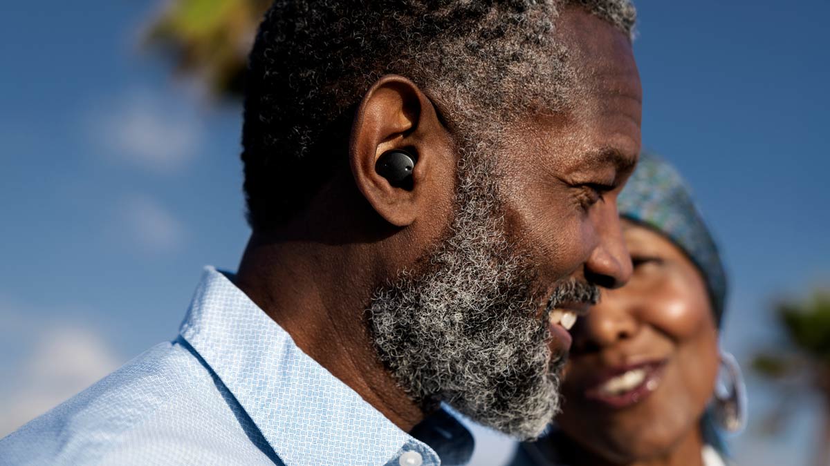 side view of person smiling while wearing black Sony hearing aid, second smiling person blurred in background