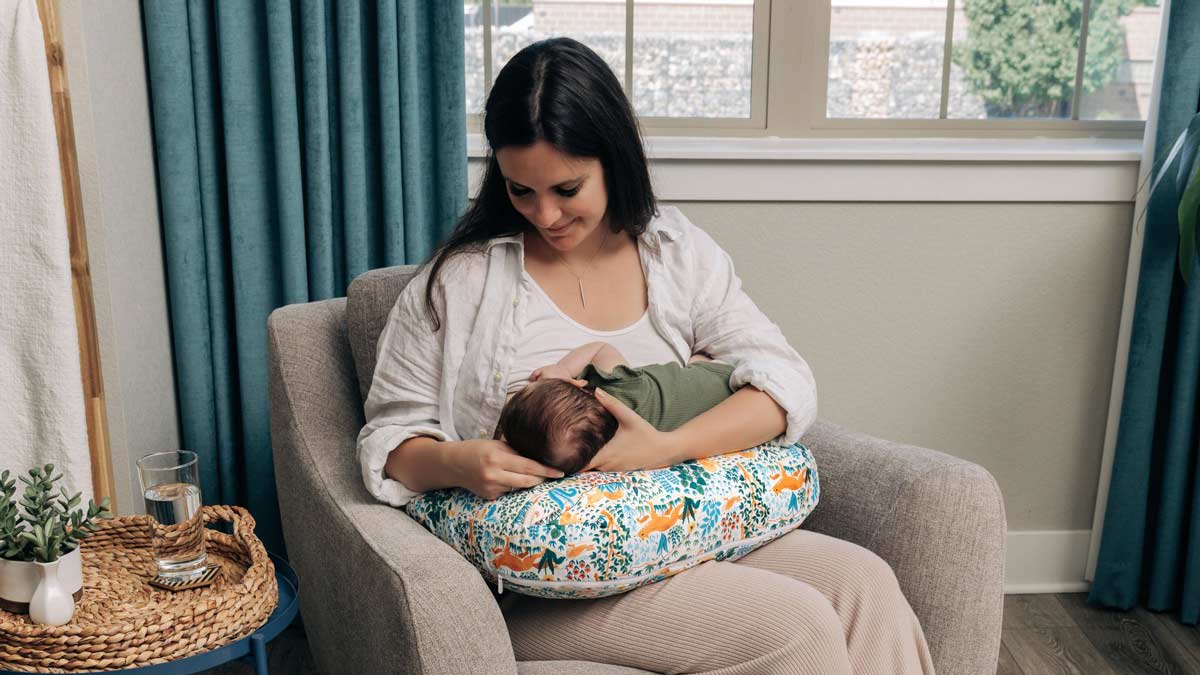 A mother sitting while breastfeeding her newborn baby with the New Boppy Nursing Pillow in Colorful Forest Animals print.