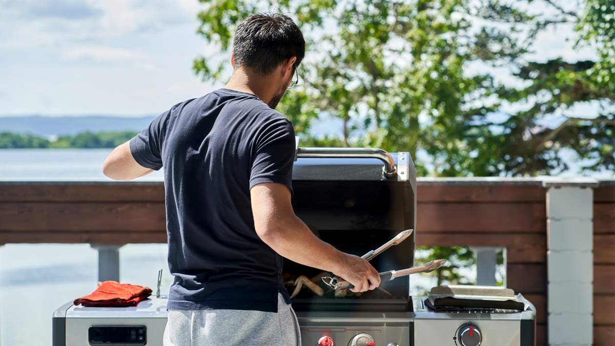 A man cooking using a grill and tongs on a lakeside balcony.