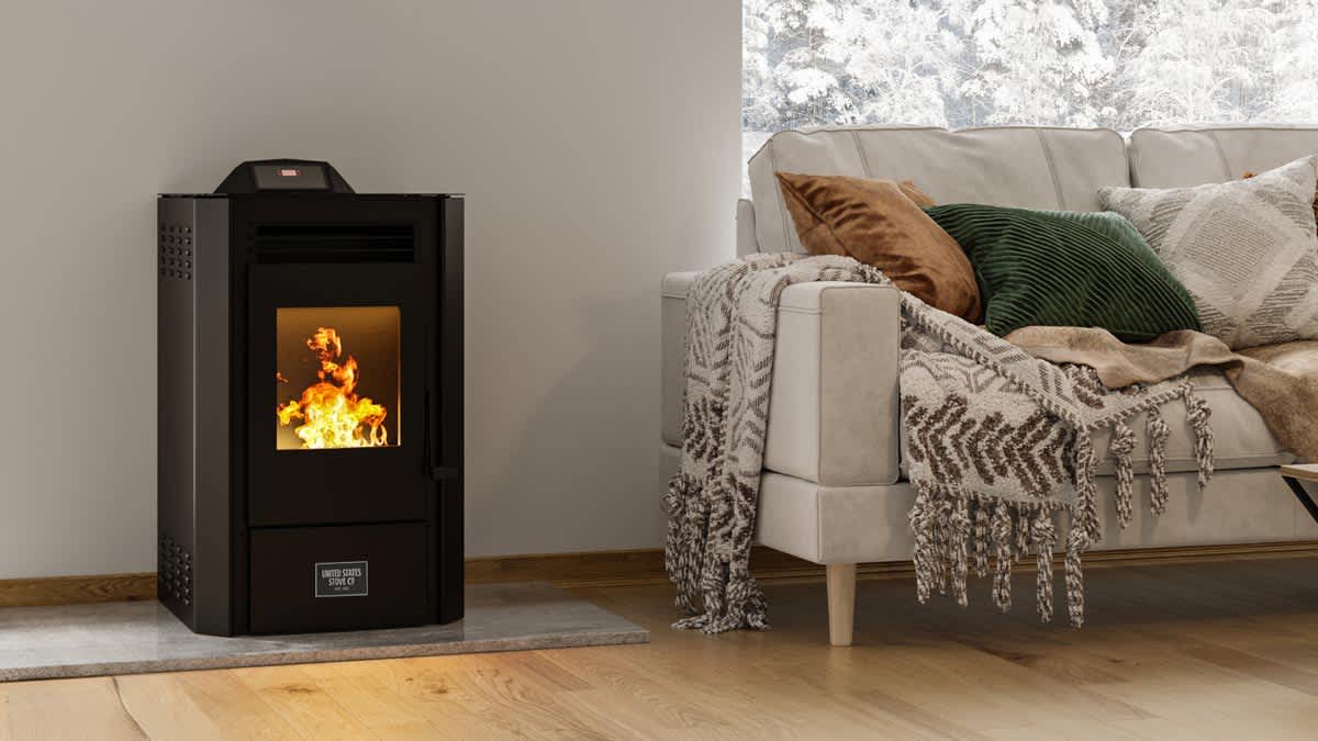 United States Stove Modern Wood Pellet Stove with 50lb Hopper in living room with beige couch with blankets and pillows, snow on trees through window behind couch