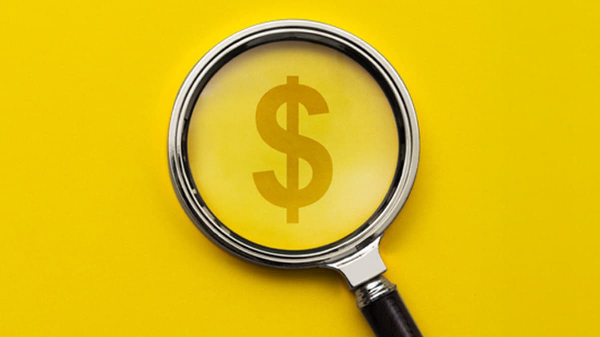 Magnifying glass examining a dollar sign on a yellow background.