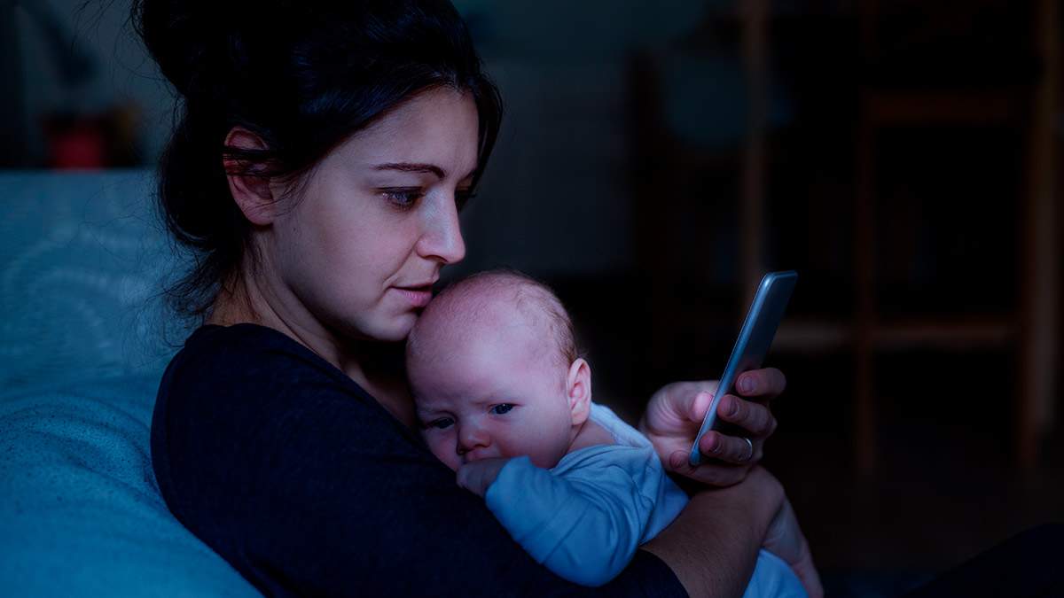 Mother holding an awake infant in the middle of the night while looking at her phone.