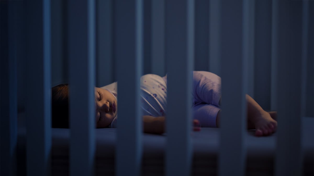 A baby seen through the bars of a crib sleeping at night.