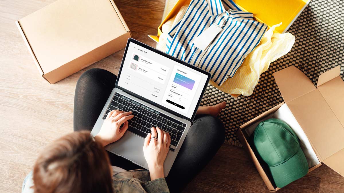A person sits on the floor using a laptop to shop online, surrounded by open boxes with clothing items including a striped shirt and a green cap.