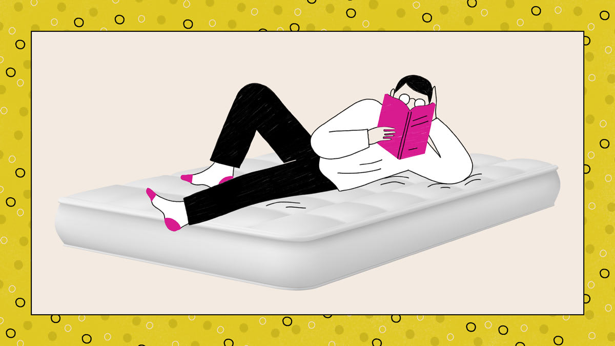 A person relaxing on a mattress, reading a book with a steaming mug nearby.